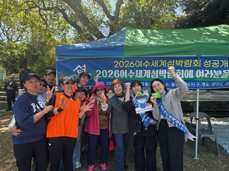 2026여수세계섬박람회 홍보부스 앞에서 주민들이 다섬이 캐릭터 손팻말을 들고 단체 사진을 찍는 모습. 야외 행사장에서 파란·초록색 천막 아래 ‘2026여수세계섬박람회 성공개최’ 현수막이 설치되어 있으며, 참가자들이 손가락 브이와 엄지 제스처로 밝게 포즈를 취하고 있다.