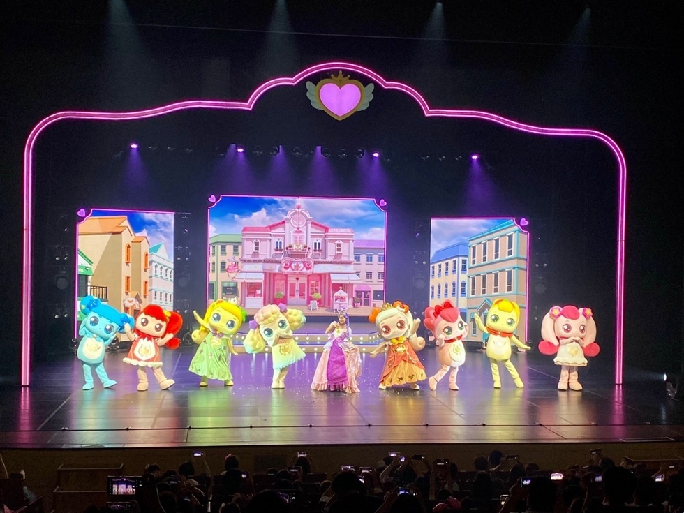 무대에서 여러 캐릭터들이 춤을 추고 있으며, 배경에는 건물들이 그려진 화면이 있다. 관객들이 공연을 관람하고 있다.