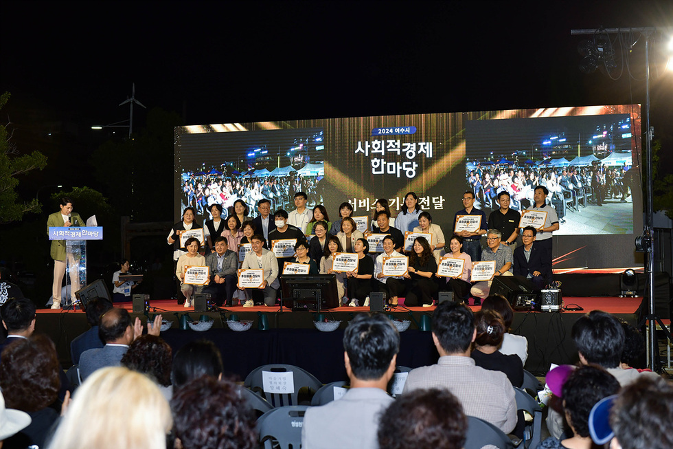 사회적경제 한마당 행사에서 여러 사람들이 무대에 모여 기념사진을 찍고 있으며, 뒤쪽 스크린에는 행사 관련 정보가 표시되어 있다.