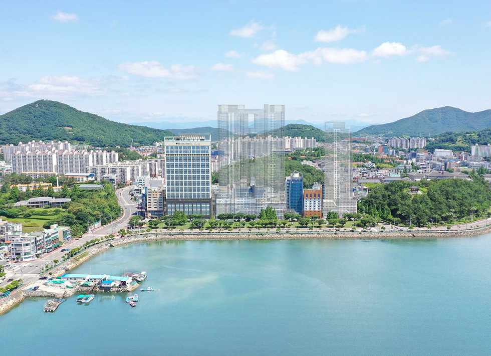 해안가에 위치한 도시 풍경으로, 여러 건물과 산이 보인다. 바다에는 작은 배들이 떠 있으며, 푸른 하늘과 구름이 어우러진 모습이다.
