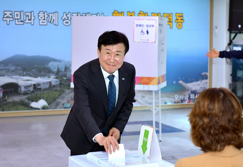 한 남성이 투표소에서 투표를 하고 있으며, 배경에는 해변과 관련된 정보가 담긴 현수막이 보인다.