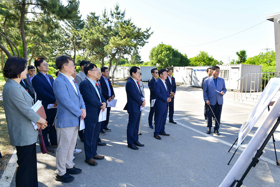 여러 명의 남성과 여성이 야외에서 발표를 듣고 있으며, 한 남성이 발표 자료를 설명하고 있다. 참석자들은 정장 차림으로 보인다.