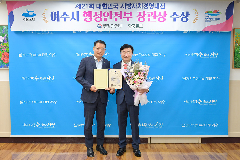 여수시장이 행정안전부 장관상 수상 후 상장과 꽃다발을 들고 기념사진을 찍고 있다. 배경에는 여수시 로고와 제21회 대한민국 지방자치경영대전 여수시 행정안전부 장관상 수상 문구가 적힌 현수막이 있다.