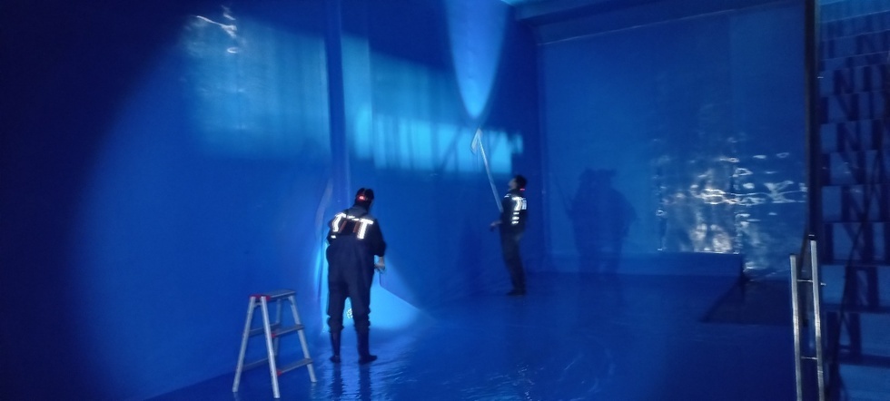 여수시 상수도 시설 내에서 작업자 2명이 파란색 방수복을 입고 대형 물탱크 내부를 청소하는 모습. 벽면은 푸른 조명이 비춰 반짝이고, 한쪽에는 사다리와 철제 계단이 보인다