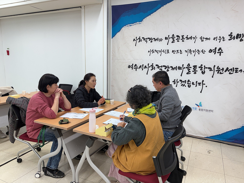 마을공동체 입문학교 2회차 과정에서 주민들이 자신들이 작성한 사업계획서를 기반으로 전문가에게 1:1 맞춤 컨설팅을 받고 있는 모습. 네 명의 참여자가 사업 계획을 보며 조언을 듣고 있으며, 배경 벽면에는 ‘여수시사회적경제마을통합지원센터’ 문구가 보인다.