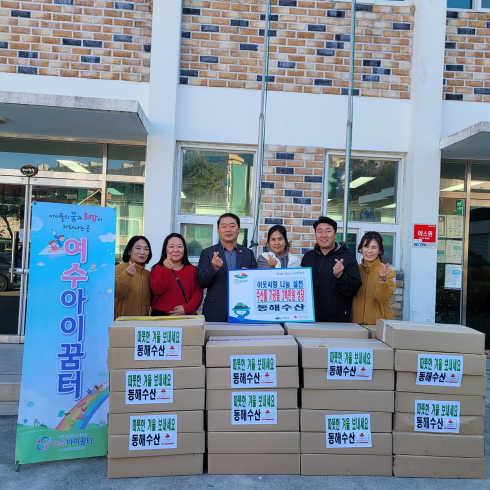 동해수산 관계자들과 여수아이꿈터 직원들이 건물 앞에서 수산물 가공품 후원 전달식을 열고 기념촬영을 하고 있다. 참가자들 뒤에는 '여수아이꿈터' 배너가 세워져 있으며, 앞쪽에는 동해수산에서 준비한 후원 물품 상자가 여러 개 쌓여 있다.